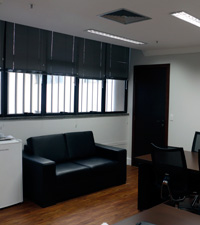 Diretoria-Geral com janelas, sof&aacute; e frigobar &agrave; esquerda, porta ao fundo e ambiente de reuni&otilde;es &agrave; direita