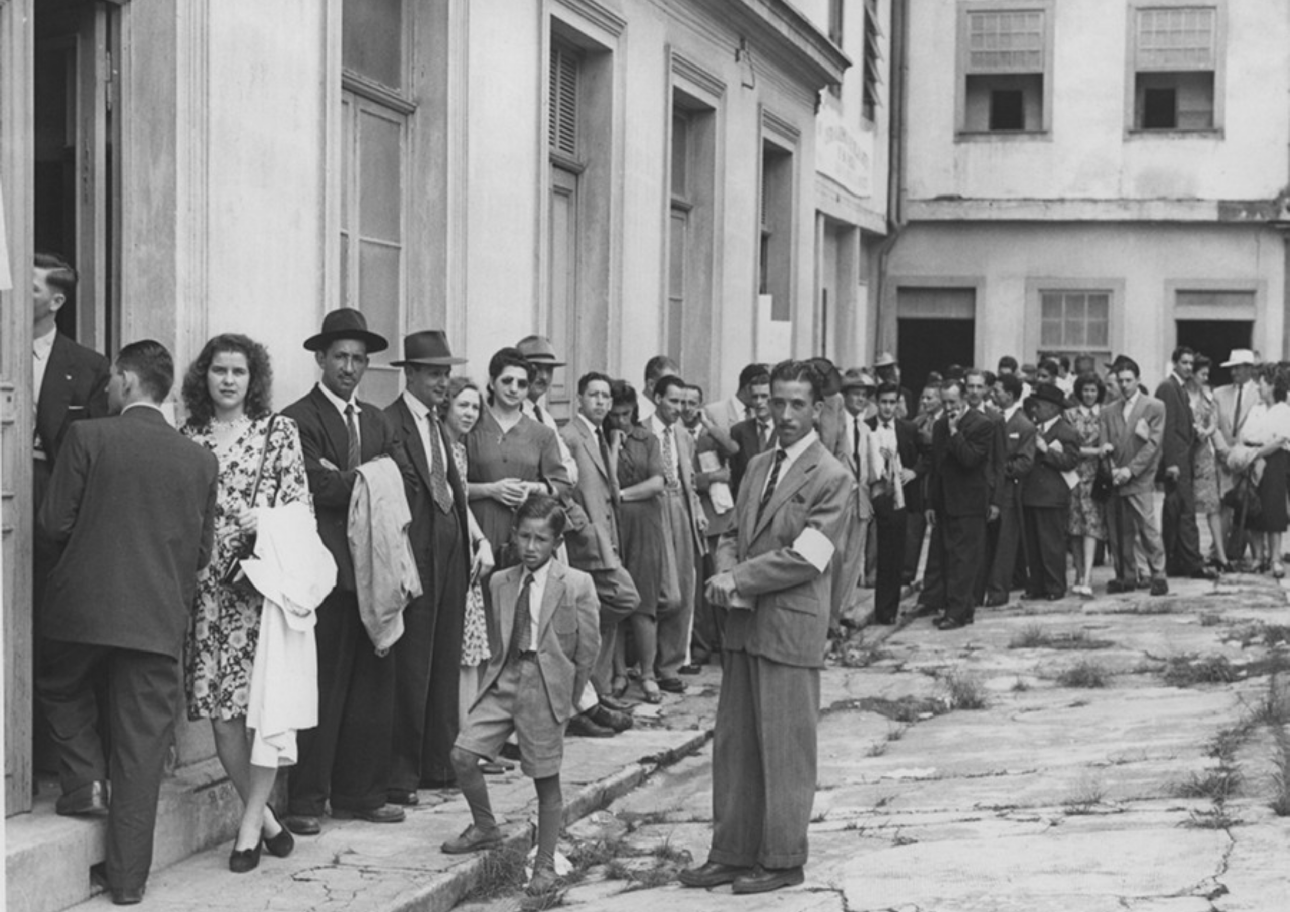 1947 - Primeira eleição direta de governadores e prefeitos após a Ditadura do Estado Novo, quando a população escolheu seus governadores e prefeitos. Fiscal observa fila de eleitores que aguardam para votar nas eleições de 1947 em São Paulo. Fonte: Estadão | Memorial da Democracia